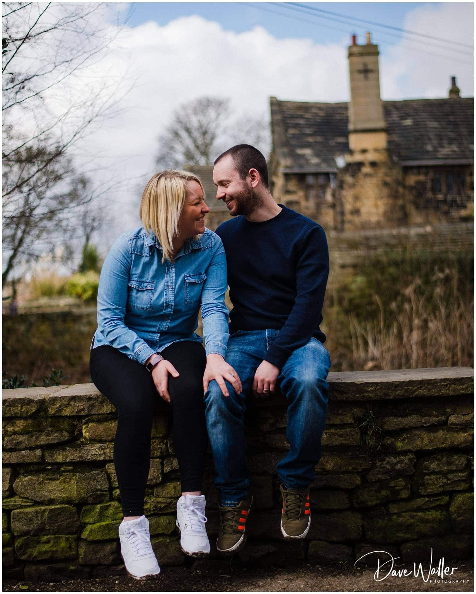 A couple sitting closely on a stone wall, smiling and looking at each other with a blurred historical building in the background during their Rachel & Danny Engagement shoot.