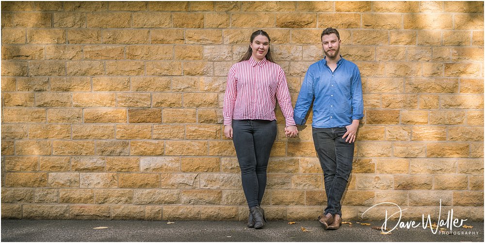 A smiling couple holding hands in front of a textured stone wall, exuding a warm and relaxed atmosphere, perfectly capturing the essence of their Autumn engagement shoot.