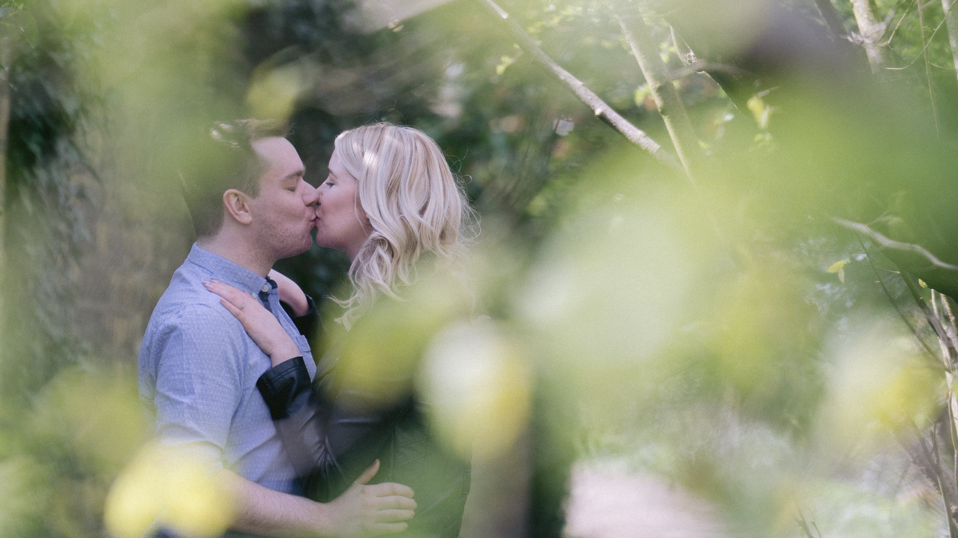 A tender moment captured among the greenery at Oakwell Hall Country Park as a couple shares a kiss, partially veiled by soft-focus foliage during their sunny couple shoot.