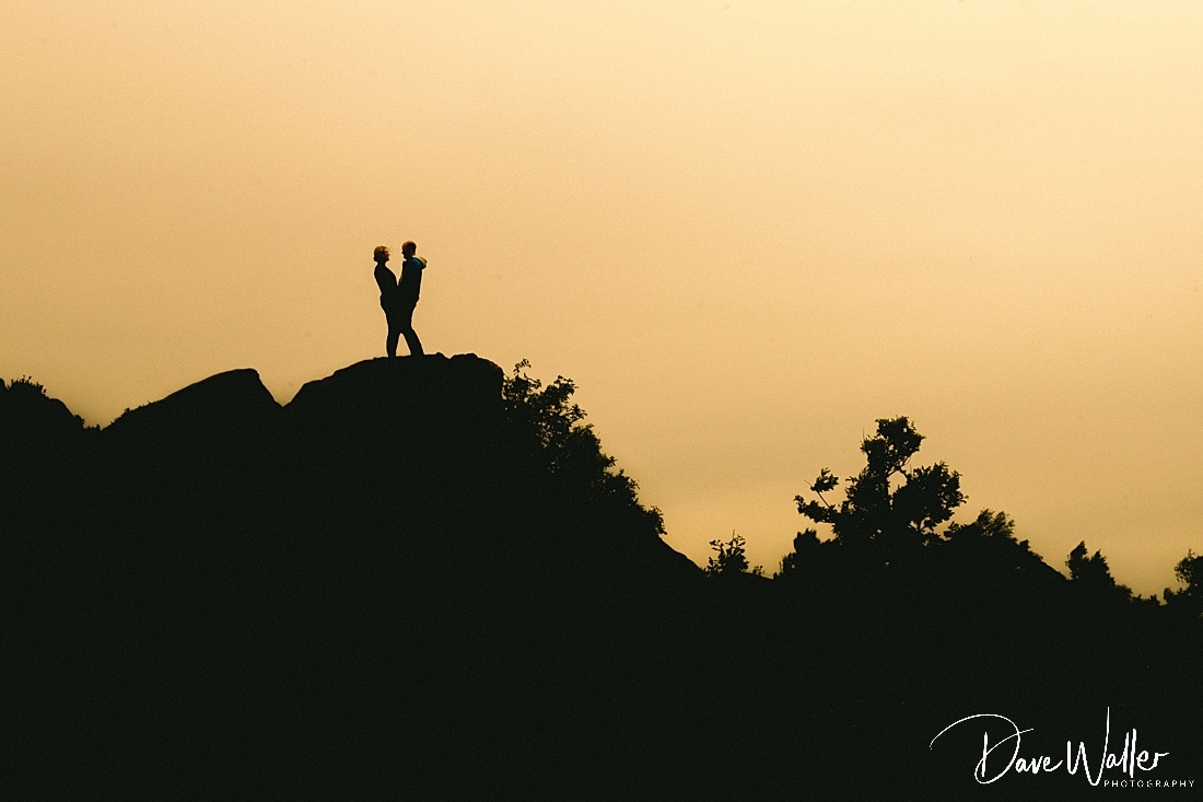 Silhouetted figures share a tender moment atop a rocky outcrop against the tranquil backdrop of a sunset sky at The Chevin Otley.