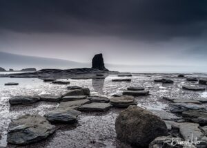 A solitary rock monolith towers majestically under a dramatic, stormy sky on a desolate beach scattered with flat stones.