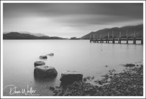 A tranquil black and white long exposure photograph capturing the serene beauty of a lake with smoothly blurred water, foreground rocks in sharp focus, and a tranquil pier extending into the distance under a motion-blurred sky