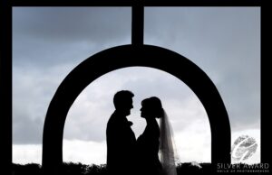 A silhouette of a bride and groom sharing an intimate moment under an archway, with a dramatic sky in the background and an emblem indicating a "silver award from the guild of photographers" on the right side.