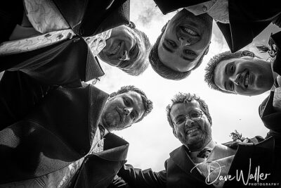A unique below-angle shot of five smiling individuals, likely groomsmen, forming a circle and looking down towards the camera on a bright day.