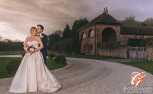 A newlywed couple sharing a tender moment, standing side by side in front of a quaint brick building under a dramatic evening sky.