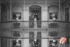 An elegant couple shares an intimate moment on the ornate balcony of a grand hall, surrounded by classical architecture and timeless charm.
