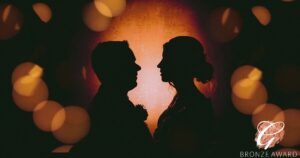 A silhouette of a couple set against a warm glowing backdrop with twinkling lights, capturing a moment of intimacy and connection, adorned with a "bronze award" seal in the corner.