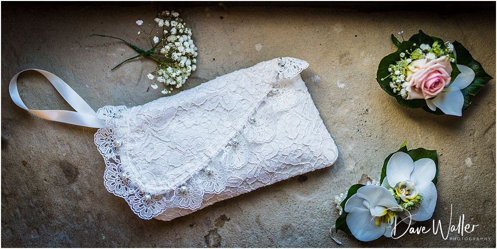 A delicate, lace-adorned bridal purse, accompanied by a boutonniere and a small floral arrangement, beautifully positioned on a rustic surface, capturing the stunning detail of a wedding day.