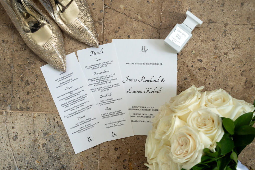 A stunning wedding invitation set graces a stone surface with white roses and gold shoes. It features an invitation, details card, and menu card for the union of James Rowland and Lauren Kelsall. Nearby, a small white perfume bottle adds an elegant touch to the detail image.