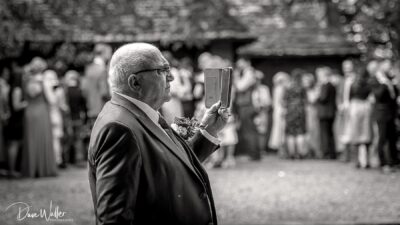A distinguished gentleman in a suit capturing a special moment with his smartphone at a social gathering, possibly a wedding, as guests in the background look on.