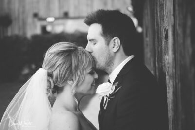 A tender moment between a bride and groom captured in a black and white photograph, as the groom softly kisses the bride's forehead, reflecting the love and intimacy of their wedding day.