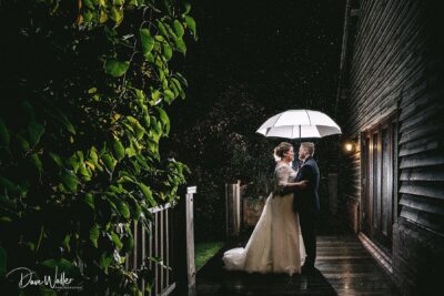 A couple shares a romantic moment under an umbrella as rain gently falls around them, illuminating their love with the warm glow of the evening lights.