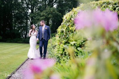 A couple on their wedding day, holding hands and walking along a garden path, surrounded by lush greenery and vibrant purple flowers.