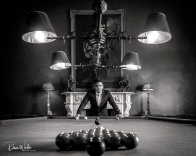 A poised player preparing for a break shot on a billiard table under a dramatic chandelier, with a confident gaze directed straight at the camera.