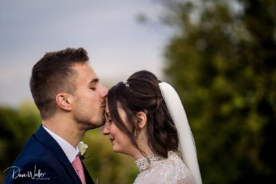 A tender moment between a newly married couple as they share a gentle kiss, with the groom's eyes closed and the bride gazing downward, surrounded by a soft natural backdrop.