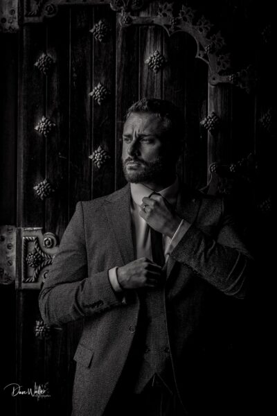 A monochrome portrait of a contemplative man in a tailored suit, poised in a moment of introspection against an ornate wooden doorway, the play of light and shadow highlighting the textures and the depth of the scene.