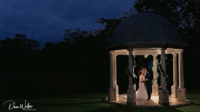 A couple shares an intimate moment within a softly-lit gazebo against the backdrop of a twilight sky.