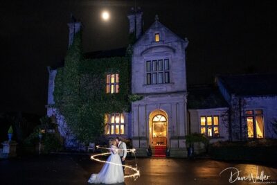 A romantic couple shares an enchanting moment with a sparkler under the glow of a full moon, in front of an elegant manor house at night.