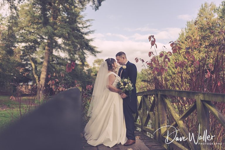 A tender moment between Sara and Scott captured on a quaint wooden bridge at Aldwark Manor amidst lush greenery and delicate autumn leaves, symbolizing the beginning of their shared journey in life.