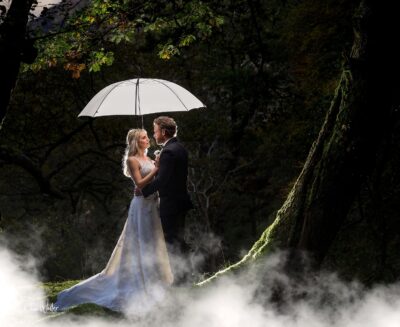 A couple shares a romantic moment under an umbrella amidst a mystical forest setting, with ethereal fog swirling at their feet and the sun filtering through the trees to illuminate them.