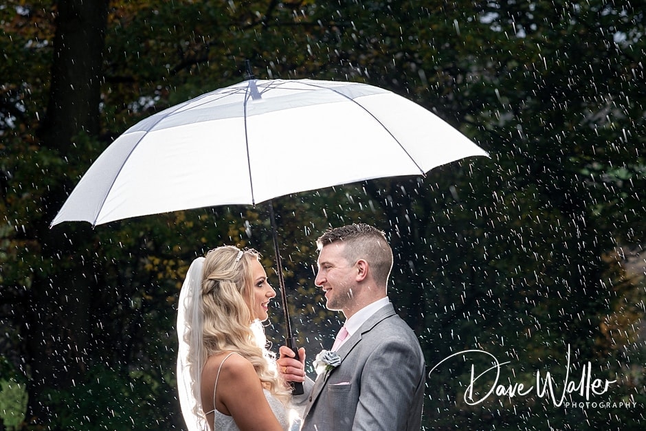 A radiant couple shares a moment of joy under an umbrella on their wedding day, with raindrops glistening around them like sparkling confetti, captured by a Leeds Wedding Photography expert.