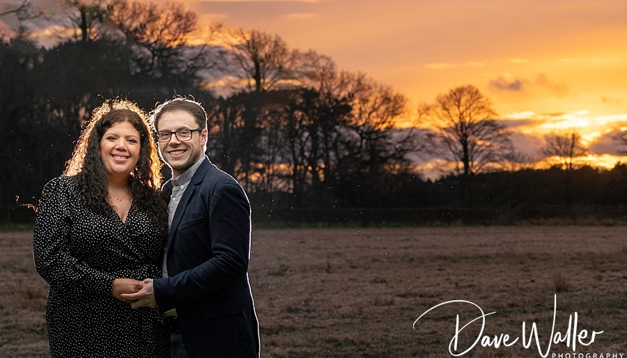 A smiling couple embracing in a field at dusk, with the warm glow of a setting sun in the background during their Couple Shoot.