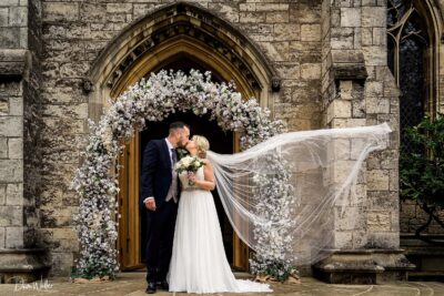 A bride and groom sharing a kiss at the church entrance, framed by an elegant floral arch and the bride’s flowing veil dancing in the breeze.