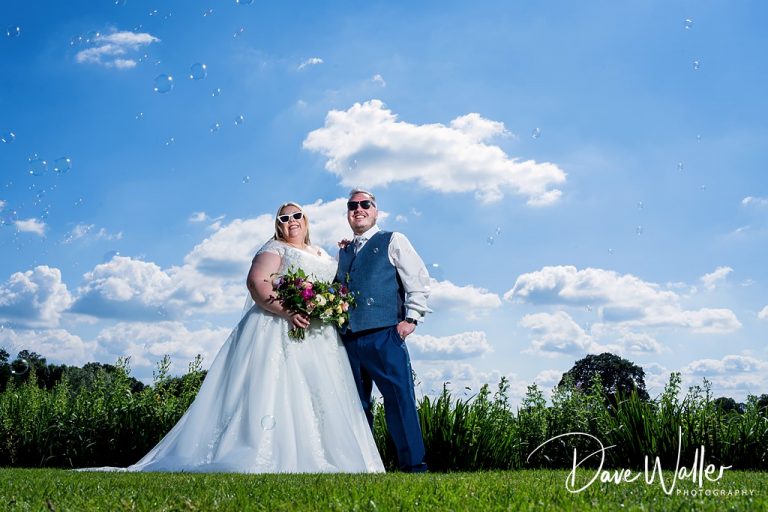 A joyful bride and groom, Katie & Pete, bask under a clear blue sky at Sandhole Oak Barn, surrounded by floating bubbles, capturing a moment of bliss on their wedding day.
