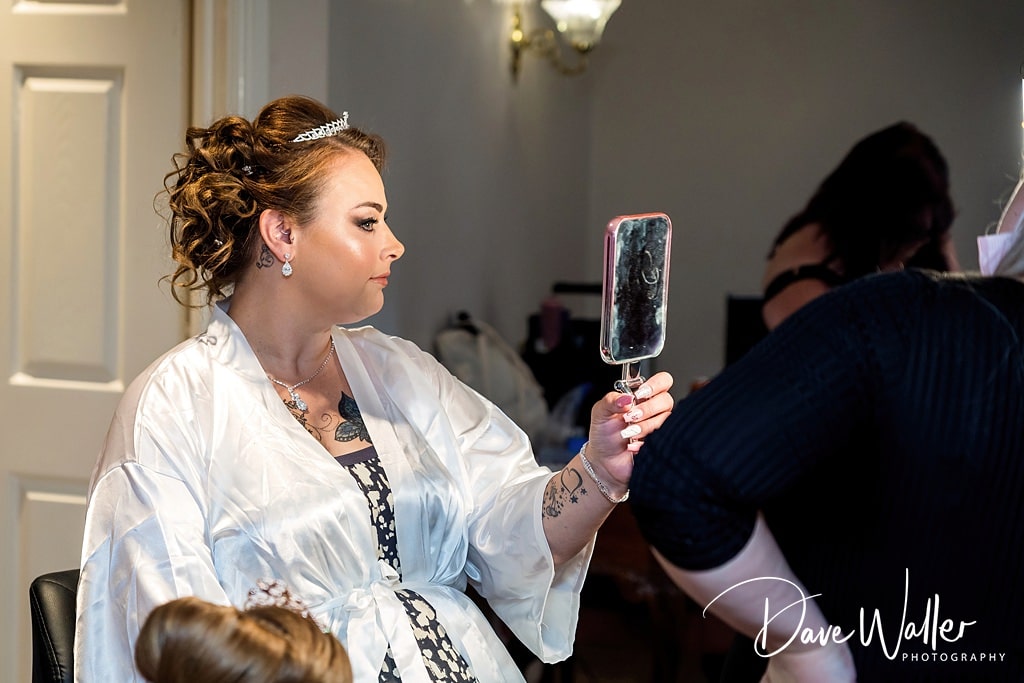 A bride, adorned with a tiara and wrapped in a white robe, gazes into a hand mirror, admiring her hairstyle as she prepares for her Waterton Park Wedding day.