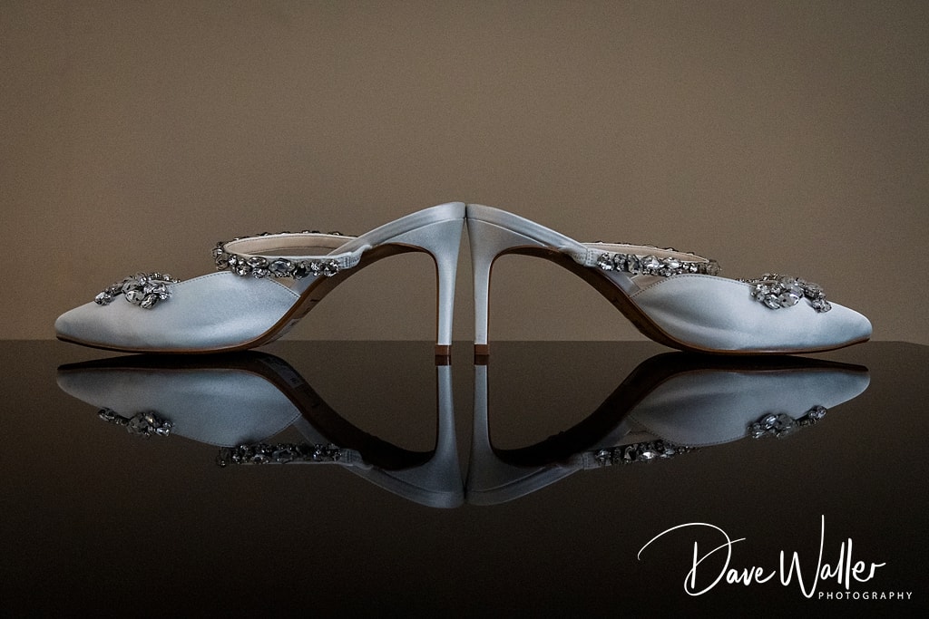 An elegant pair of bridal shoes with sparkling embellishments, poised symmetrically on a reflective surface at Hollins Hall Hotel for Sophie & Nicholas's Wedding.
