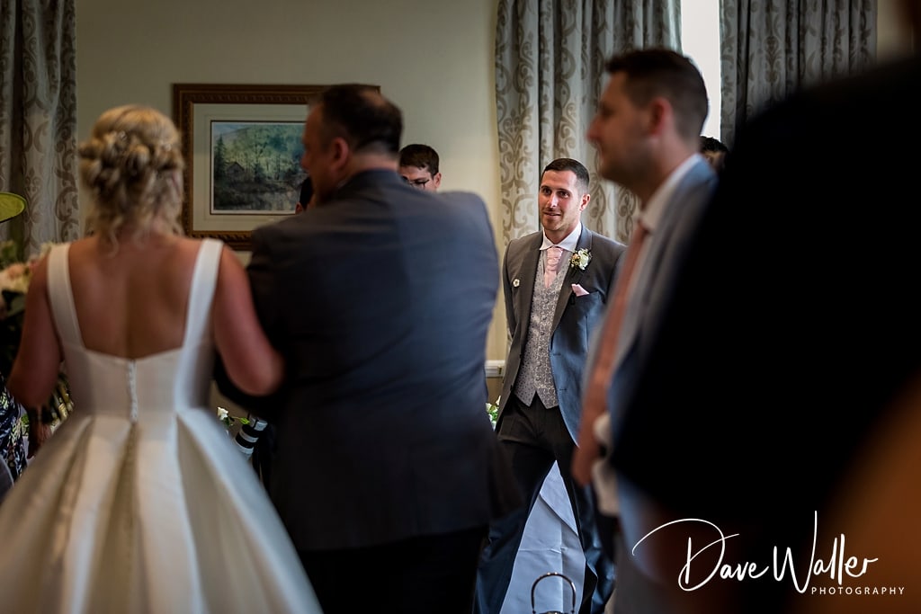 A poignant moment during Sophie & Nicholas's wedding ceremony at Hollins Hall Hotel captured from a unique perspective, with the groom awaiting expectantly as the bride approaches, surrounded by guests.