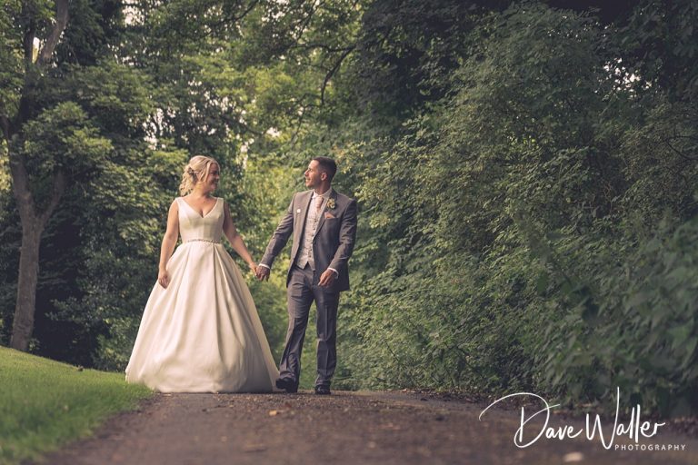 Sophie & Nicholas, in wedding attire, holding hands and walking down a serene tree-lined pathway at Hollins Hall Hotel, exchanging loving glances.