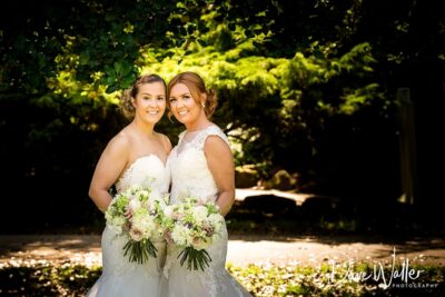 Ella & Sophie, two smiling brides in elegant wedding dresses, are posing together on a sunny day, each holding a beautiful bouquet of flowers.