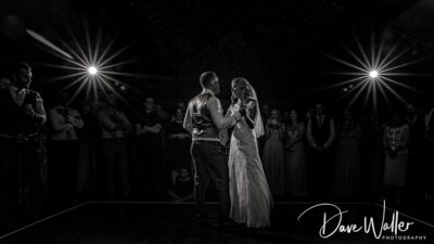 A spotlight moment of affection as Rebecca & Jonathan shares their first dance, surrounded by friends and family, captured in a timeless black and white photograph.