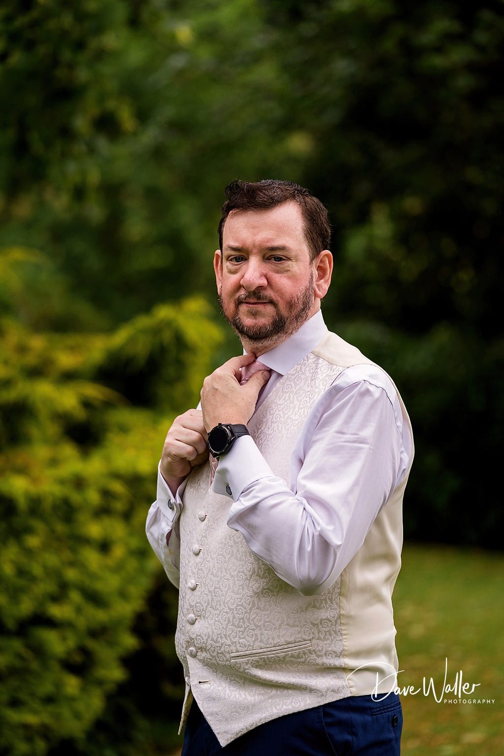 A well-dressed man adjusting his tie outdoors with a thoughtful expression, captured by Sanchia and James, the Wedding Photographer at Waterton Park.