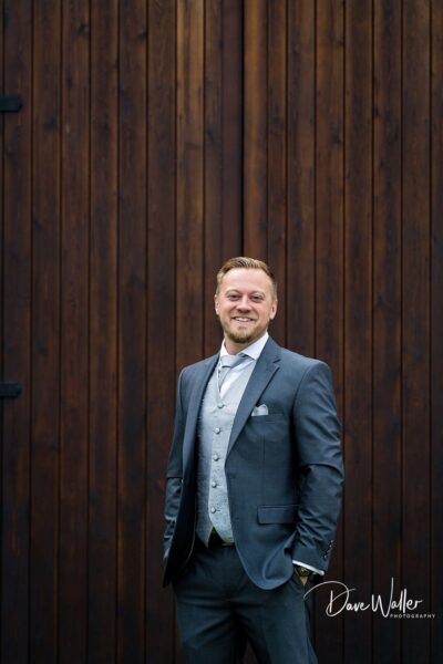Elegantly dressed man posing with confidence against a rustic wooden backdrop for Normans Wedding Photography.