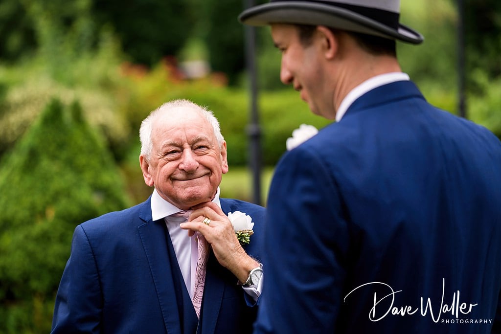 A cheerful elderly gentleman in a suit shares a heartfelt moment with a younger man in a blue suit and fedora, who seems to be adjusting the elder's boutonniere. This touching scene,