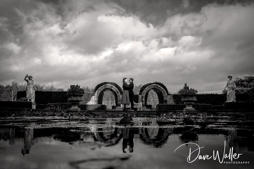 A dramatic black and white couple photography capturing a romantic duo posing between curved stone structures at Allerton Castle, with their reflection mirrored in the water, while two photographers capture the moment from different angles under a