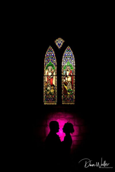 A couple stands in contemplation before the vibrant hues of a stained glass window, their silhouettes framed in the peaceful glow of a sacred space.