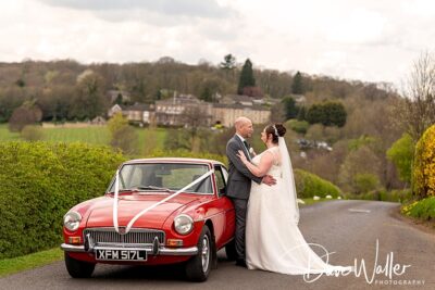 A loving couple sharing a tender moment on a scenic road beside a classic red car, with the lush countryside stretching out behind them.