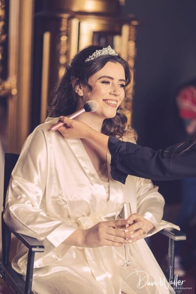A radiant bride-to-be in a silk robe, wearing a tiara and smiling joyfully, holds a glass of champagne while getting her makeup done before the wedding ceremony.