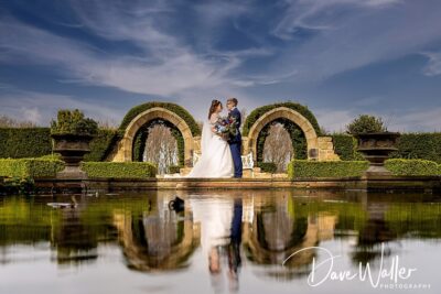 A couple in wedding attire sharing an intimate moment by a reflection pool, with arching hedges and a clear blue sky in the background.