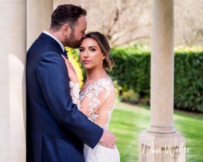 A tender moment between a groom in a dark suit and a bride in a lace dress, sharing an affectionate embrace near classic columns in a serene garden setting.