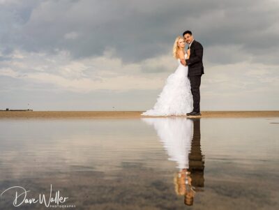 A newlywed couple embracing on a serene beach, with their affectionate pose mirrored in the water beneath a cloud-adorned sky.
