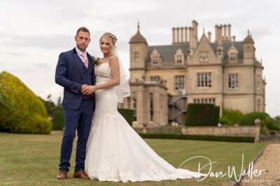 A newlywed couple holding hands and posing for a photograph in front of an elegant manor house.