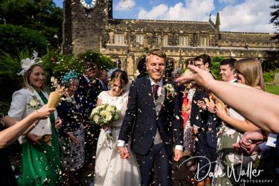 Joyful newlyweds walking through a shower of confetti with friends and family celebrating around them, against the backdrop of a picturesque historical building on a sunny day.