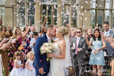 A joyful newlywed couple shares a kiss amidst a shower of confetti, surrounded by smiling friends and family celebrating the festive occasion.