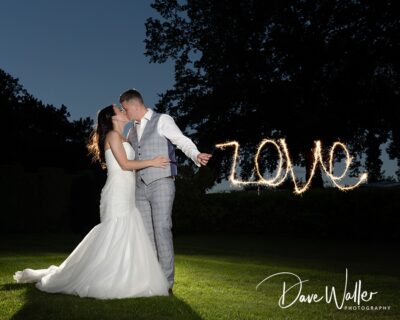 A radiant couple shares a kiss on their wedding night, illuminated by the timeless word "love" written in sparkling light against the evening backdrop.