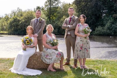 A joyful bridal party posing outdoors on a sunny day, with the bride sitting on a hay bale and attendants standing around her, all smiling and holding bouquets against a backdrop of a tranquil pond.