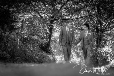 A couple walks hand in hand down a sun-dappled pathway surrounded by lush greenery, sharing a moment of joy and connection in their elegant suits.